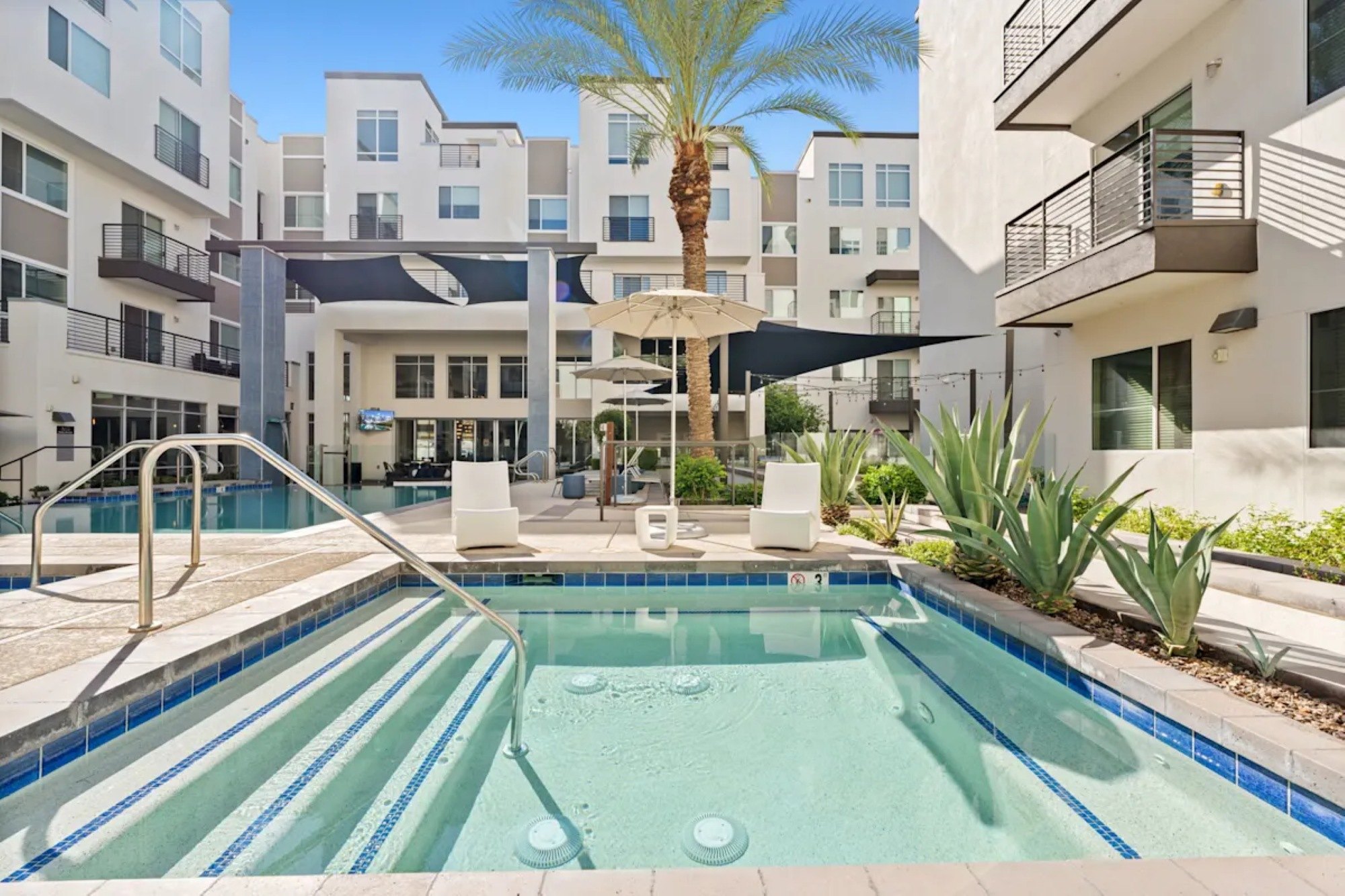 An outdoor hot tub and swimming pool surrounded by white modern apartment buildings, lounge chairs, and palm trees.